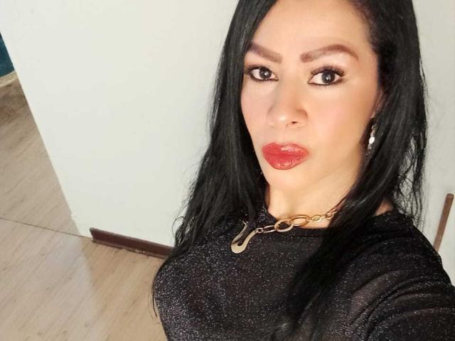 SofiMontes with long black hair, red lipstick, and a glittery black top, wearing a gold chain necklace, has her tits showing through the top, and a serious expression.