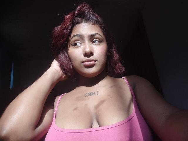 AmberQueeny with dark red hair, pink tank top, and "SEEL" tattoo on chest, gazing sideways, sunlight on face, hand in hair, dark background, slightly shadowed.