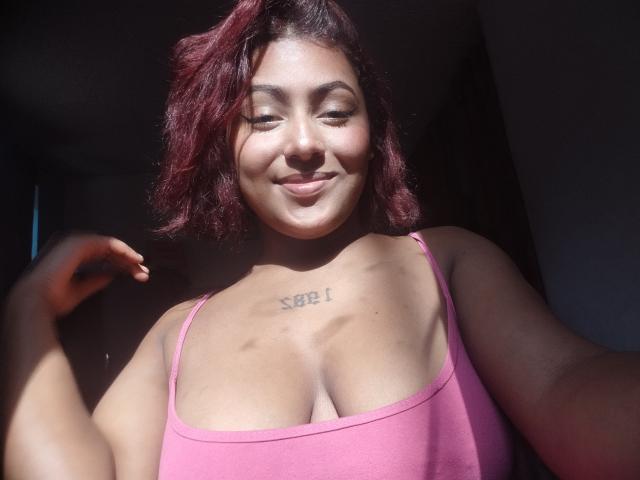AmberQueeny with red hair, smiling, wearing a pink top, showing her tits, tattoo above her cleavage, sunlit, hand raised.