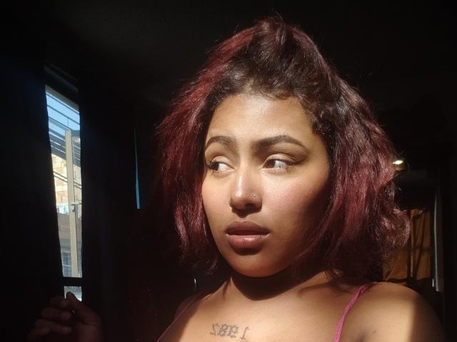AmberQueeny with dark red, wavy hair, brown skin, and a nose piercing, looking to the side, sunlight on her face, small tattoo above her tits, wearing a pink top.