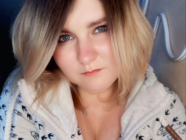 MinistraX has blonde, shoulder-length hair, blue eyes, and a slightly flushed face. She's wearing a white, patterned hoodie with a small tattoo near her collarbone.
