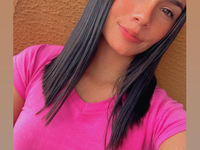 KimmyK met recht zwart haar, een roze shirt en een lichte glimlach, die haar borsten en zachte, roze lippen laat zien, voor een bruine muur met een structuur..