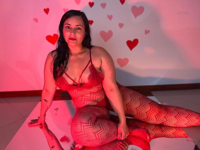 MichellMonet with wet black hair, red zigzag fishnet bodysuit, sitting on white floor, legs crossed, showing her tits and pussy, red hearts on white wall, red lighting.