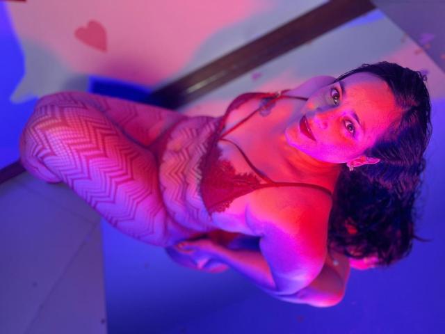 MichellMonet with curly brown hair, red lace bra, and zigzag-patterned fishnet leggings, kneeling, looking up, lit by pink and blue lights, showing her tits and ass.