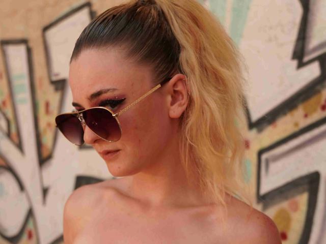 Melly4you-hot with blonde ponytail, wearing aviator sunglasses, and no shirt, showing her bare shoulders and a slight smile, looking to the side.