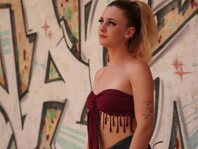 Melly4you-hot with blonde ponytail, wearing a maroon strapless top with fringes, exposing her tits, tattoo on right arm, standing against graffiti, looking to the side with a slight smile.