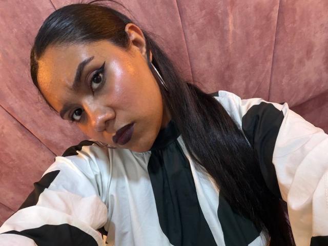 SashaSpicy has shiny brown skin, dark purple lipstick, and black eyeliner. She's wearing a black and white striped shirt, long black hair, and silver hoop earrings.