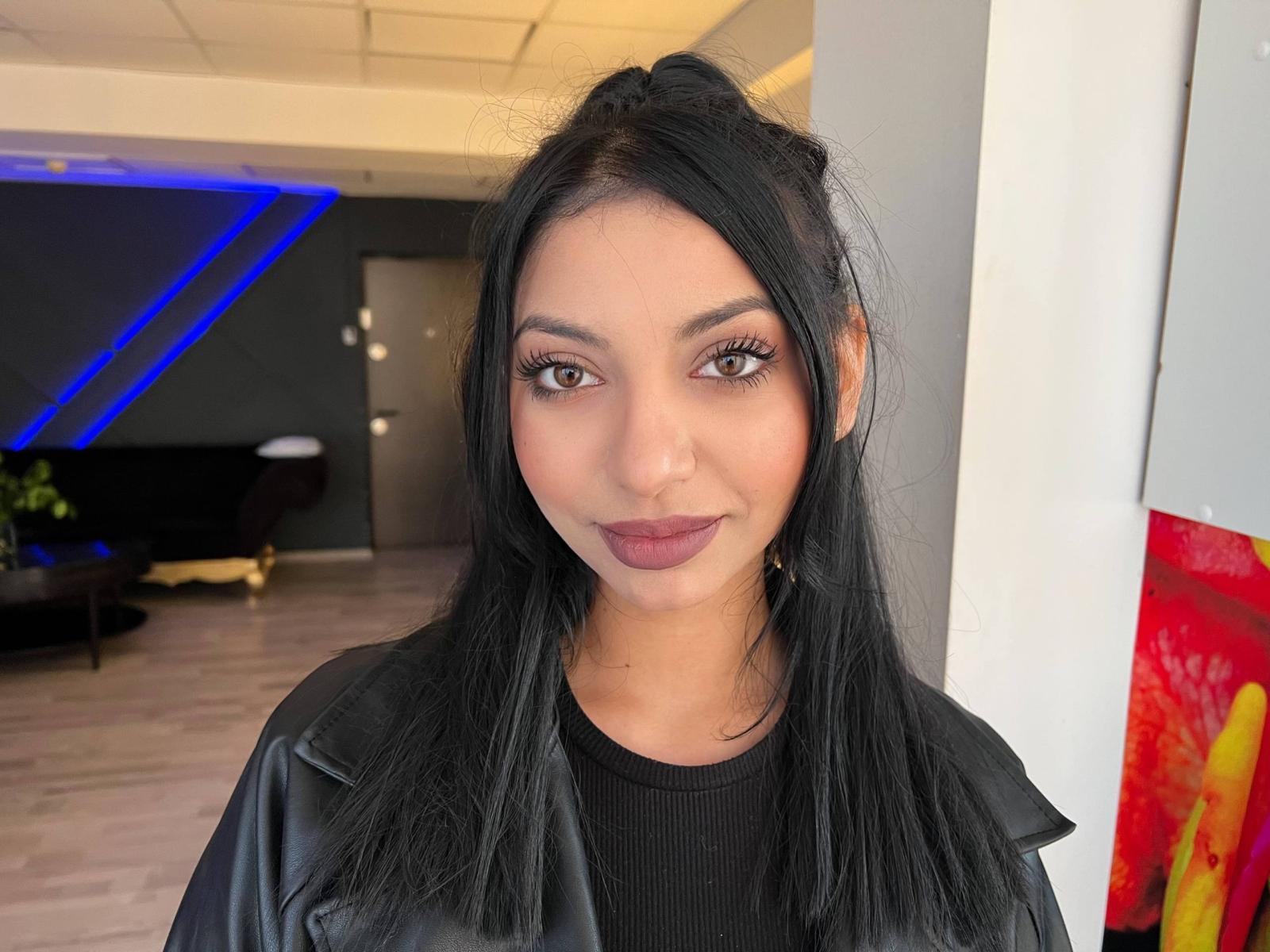 NyxAngel with long black hair, brown eyes, and dark purple lipstick, wearing a black leather jacket, smiling softly, standing in a modern room with blue and white accents.