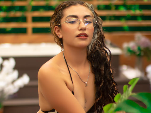 AdaraAngel with wet curly brown hair, glasses, and black halter top, showing her tits, has a necklace, and a confident, sexy look, in a colorful, blurred background.
