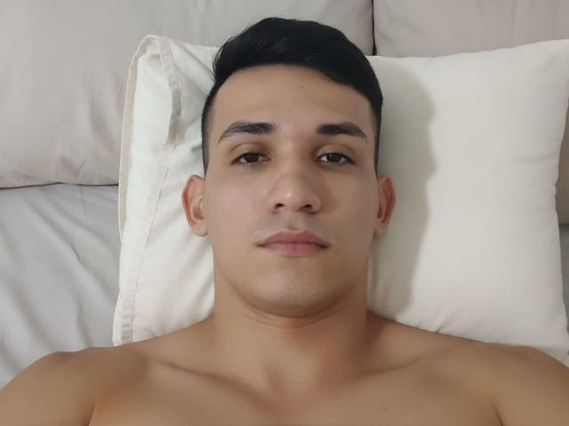 AronRieff, shirtless, lying on white pillow, black short hair, brown eyes, neutral expression, slight tan, small nose, short upper lip, bare shoulders, head centered in image.