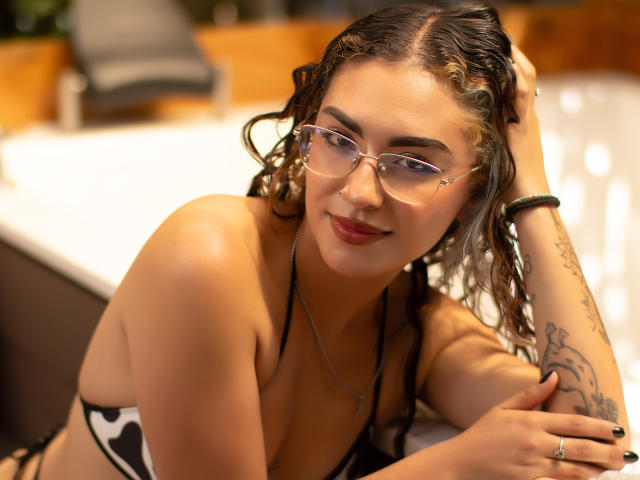AdaraAngel with curly brown hair, glasses, and a black-and-white bikini, smiling at the camera, showing her tits and tattooed arm, wearing a black bracelet, in a sunlit room.