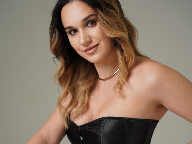 Portrait of JenifferBrown with light brown skin, wavy blonde and dark brown hair, and subtle makeup. She has dark eyebrows, brown eyes, and a small, closed-lip smile. She's wearing a strapless black leather corset that highlights her medium-sized breasts. A simple gold necklace rests on her chest. The background is a plain, light grey. Her hair is styled in loose waves, and she has a few small freckles on her shoulders. The image is sharply focused, with soft lighting that emphasizes her natural beauty.