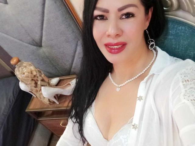 SofiMontes, con lunghi capelli neri, rossetto rosso, collana di perle, abito bianco in pizzo e orecchini, tiene in mano un flacone di profumo di vetro, sorride alla fotocamera, mostrando il seno..