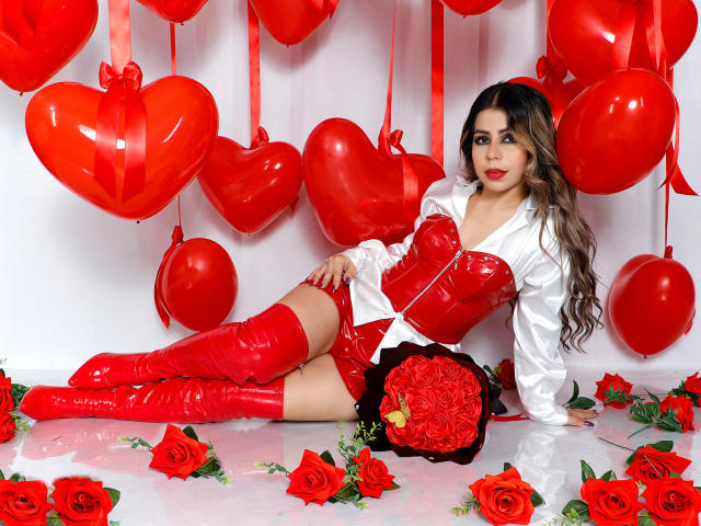 NicolBennett with red latex corset, white shirt, red thigh-high boots, red heart balloons, and red roses, holding a red rose bouquet, laying on white floor.