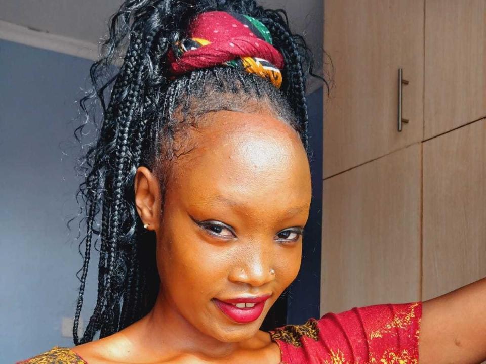 SugarBelly has dark brown skin, red lipstick, and long black braids in a high ponytail with a red and yellow patterned scrunchie. She's wearing a red dress with gold accents.