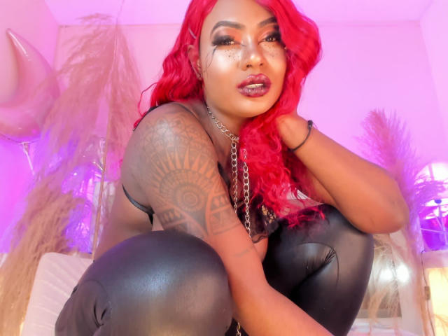 PaulinaRuiz with bright red hair, black leather pants, tattooed arm, and chain necklace, crouching, showing her shiny ass, looking at the camera with glossy lips.