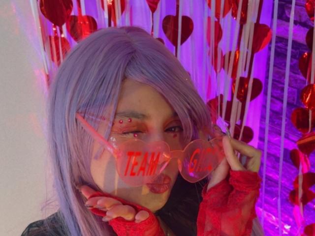 YukkyKuran with purple hair, wearing red heart-shaped glasses, red fingerless gloves, and red face piercings, holding glasses with red text, in a colorful, heart-themed setup.
