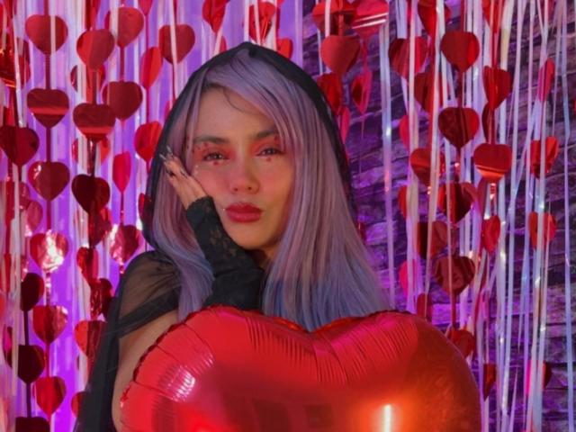 YukkyKuran with purple hair, black hood, and red lipstick holding a shiny red heart balloon, wearing a black lace sleeve. Boobs partially visible, cute and sexy.