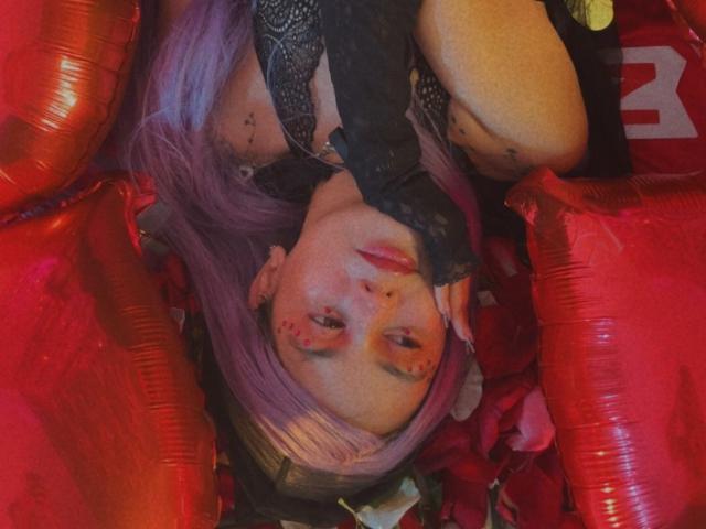 YukkyKuran with purple hair, black lace top, and gloves, surrounded by red balloons, looking up with red makeup and tattoos. Boobs partially visible, face centered.