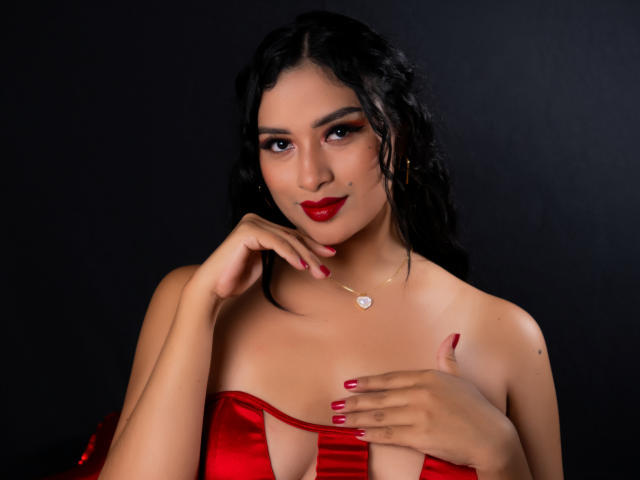 AdalinaVera with black wavy hair, red lipstick, and red nails, wearing a red satin top, touching her tits, hand on chest, necklace, dark background.