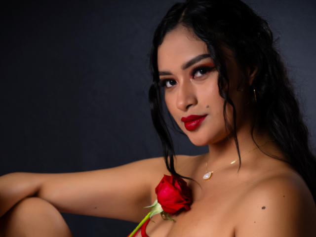 AdalinaVera with long black hair, red lipstick, and dark eye makeup, holding a red rose against her bare, medium-sized tits, wearing a small gold necklace against a dark background.
