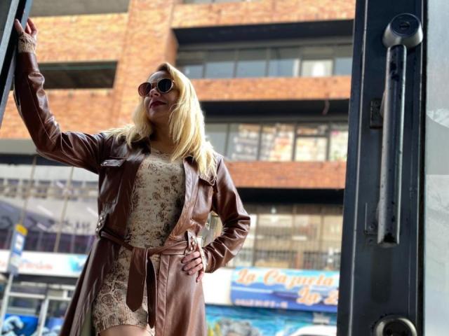 SamanthaJoneX with blonde hair, wearing a brown leather jacket and lace dress, standing in a doorway, hands on hips, sunglasses on, looking confident, sunlight on her face.
