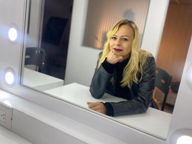 SamanthaJoneX with blonde hair, red lipstick, and a dark leather jacket, leaning on a white counter, hand under chin, in front of a mirror with light bulbs.