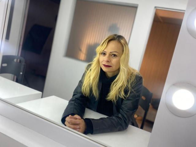 SamanthaJoneX with blonde hair, black jacket, and red lipstick, leaning on a white counter, hands clasped, smiling subtly, in a brightly lit room with a circular light.