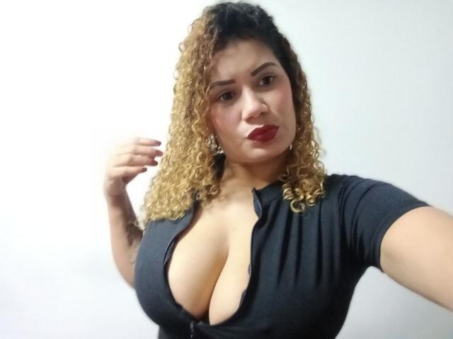 ZoeWeber ha i capelli biondi e ricci, un seno prosperoso e un profondo scollatura in una maglia nera. Guarda la fotocamera con il rossetto rosso e un leggero sorriso..