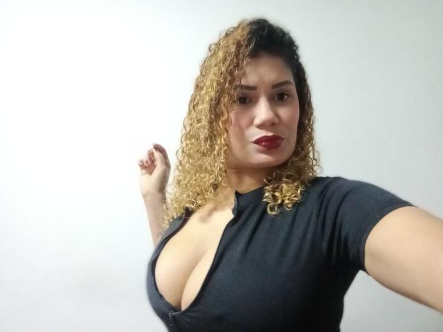 Zoe Weber, con i capelli biondi ricci, il rossetto rosso e un profondo scollo in una maglia nera, alza il braccio destro. Ha un'espressione sicura, leggermente compiaciuta..