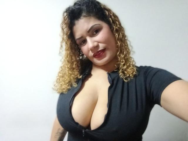 ZoeWeber tem cabelos loiros cacheados, sobrancelhas escuras e batom vermelho. Ela está vestindo uma blusa preta com zíper e um decote profundo, que mostra seus seios grandes. Ela está sorrindo para a câmera..