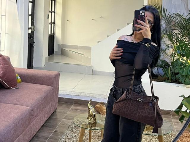 NyraVanel with black off-shoulder top, black pants, leopard purse, holding phone, covering boobs, long black hair, brown sofa, glass table, stairs, and plants in background.
