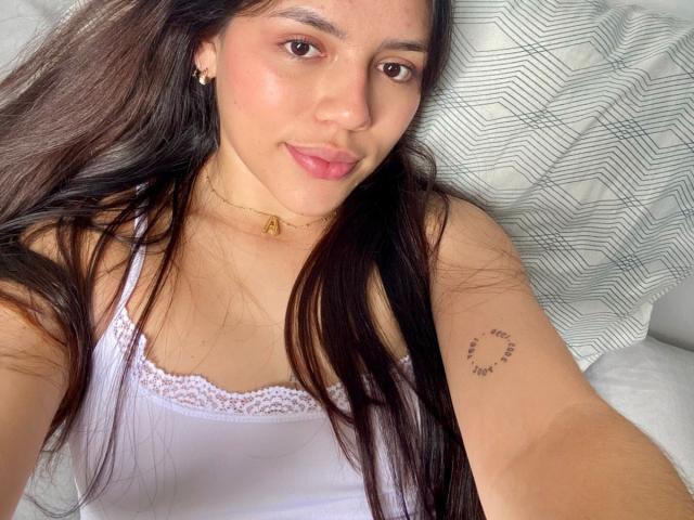 SophiaMarx has long brown hair, pink lips, and a small tattoo on her right arm. She's wearing a white lace-trimmed tank top and a gold necklace.