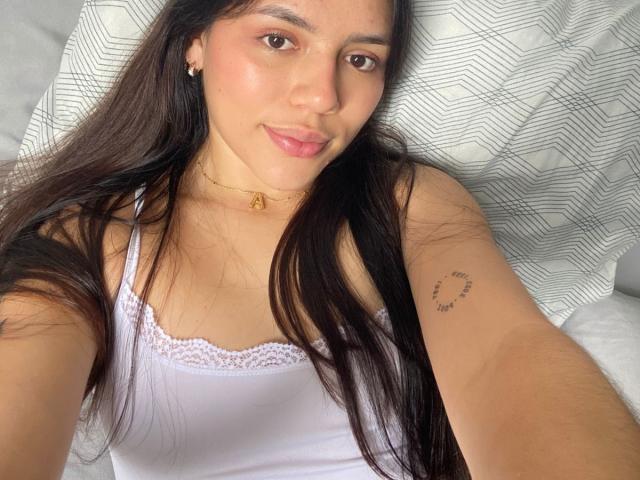 SophiaMarx with long dark hair, pinkish-brown skin, and small tits, wearing a white lace-trimmed tank top, lying down, smiling, gold necklace, small tattoo on right arm.