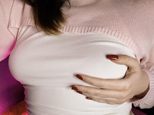 Close-up of Rubbi's chest and hand. Rubbi wears a pink ribbed sweater over a white tight-fitting top. Rubbi's right hand, with red painted nails, is placed over her large, rounded breast, slightly lifting the white top. The texture of the sweater is ribbed, while the white top is smooth. The image focuses on the contrast between the pink sweater and the white top. The skin is light, and the hand is visible with fingers spread. The lighting is even, highlighting the textures and colors.