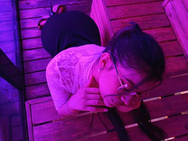 Adaallen, with glasses and braided black hair, is on her knees, wearing a white lace top and black skirt, under pink and purple lighting, touching her chin.
