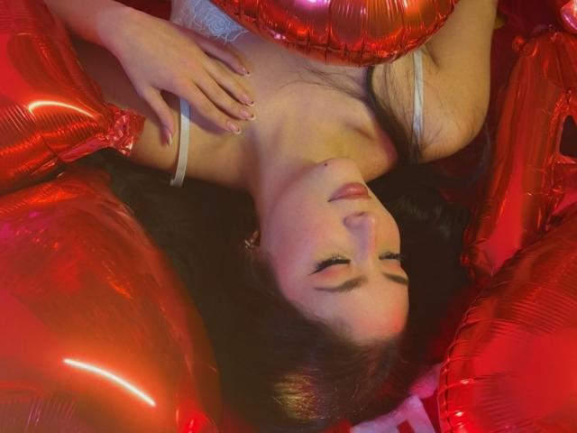 StarDustt's face, eyes closed, surrounded by shiny red balloons, lying upside down, with one hand on her chest, wearing a white strap top, looking relaxed and content.
