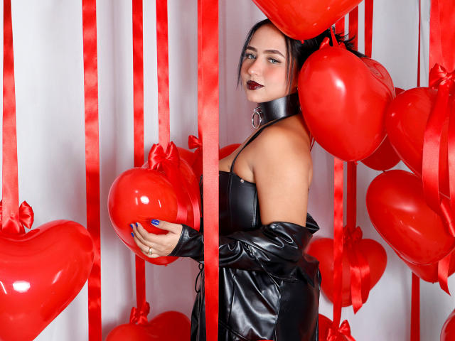 SophiaBlakes with red heart balloons, wearing black leather off-shoulder top, choker, dark lipstick, holding heart balloon, red ribbons, blue nails, looking over shoulder, medium tits, fair skin.