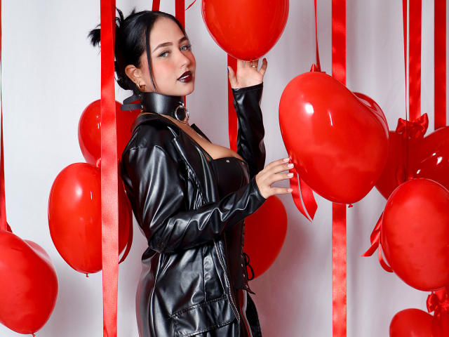 SophiaBlakes with black leather jacket, red balloons, and dark lipstick, touching a balloon with her right hand, showing her tits slightly, white background, red ribbons, black hair up.