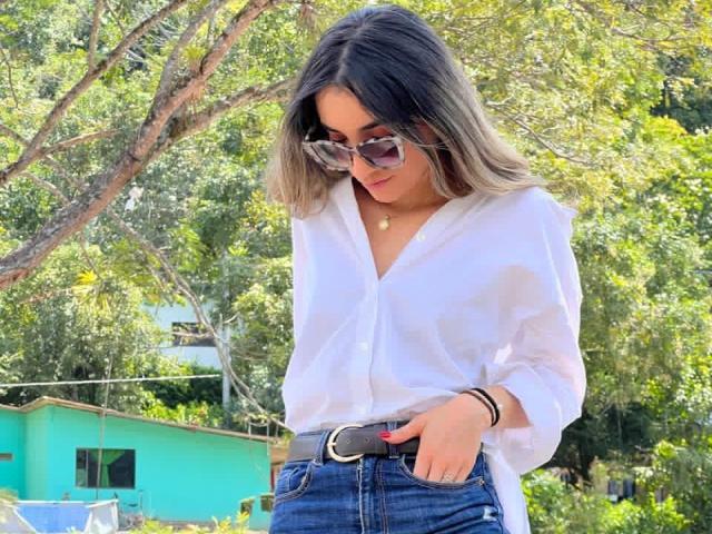 AdharaCooperr stands outdoors, wearing a white, long-sleeved button-up shirt with the top button undone, and blue jeans with a black belt. She has long, straight, ombre hair and wears dark sunglasses. Her right hand is in her jeans pocket, with a black bracelet on her left wrist. She looks down slightly, with a subtle smile. The background shows green foliage and a turquoise house. The composition is balanced with natural light highlighting her face and hair.