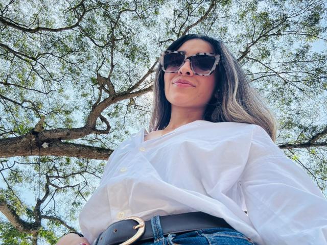 AdharaCooperr with large tits, wearing a white button-up shirt and dark sunglasses, standing under a tree, looking down at the camera with a slight smile.