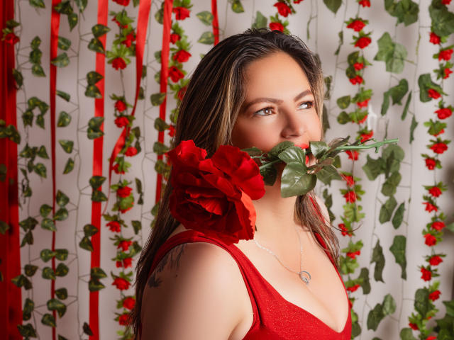 LadyLure com uma rosa vermelha na boca, segurando-a perto de seus seios fartos, vestindo um vestido vermelho, um colar de prata, cabelos castanhos e olhos verdes, com uma expressão sedutora..