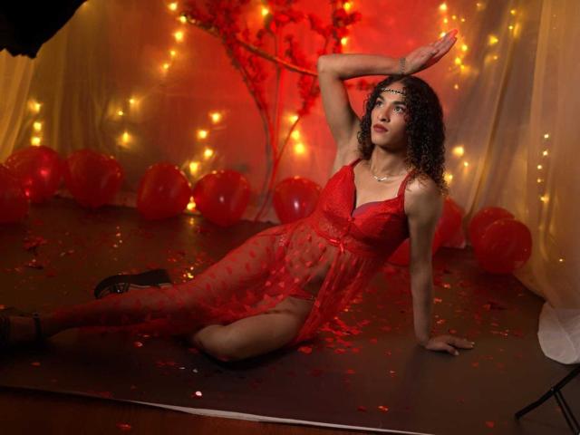 StefannyMia, with curly dark hair, wears a sheer red lace dress, exposing her small breasts and red panties. She's reclined on a stage, one arm raised, the other resting on the floor. Red petals and balloons surround her, and warm fairy lights illuminate the background. Her skin is light brown, and she has a confident, seductive expression. The image is rich in red and warm light, emphasizing a sensual, theatrical atmosphere.