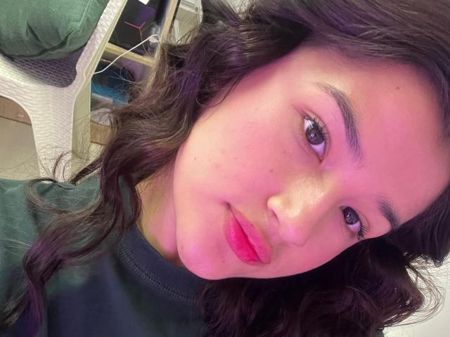 Iris Blair, con capelli neri ondulati, pelle leggermente rosata, rossetto rosso e occhi verdi, indossa un maglione verde scuro e guarda leggermente a sinistra in un selfie..