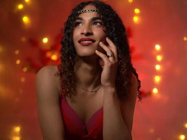StefannyMia with curly brown hair, red dress, and headband, smiling, hand near face, wearing ring, soft red and yellow bokeh lights in background.