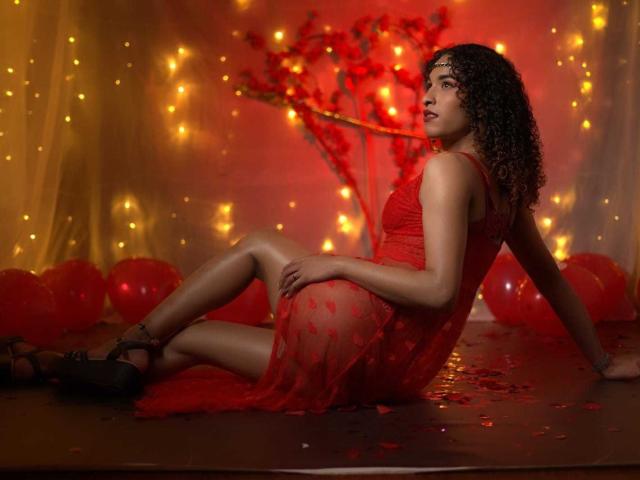 StefannyMia, with curly brown hair, wearing a red lace dress, sits on the floor, legs bent, showing her ass, surrounded by red balloons and warm fairy lights.