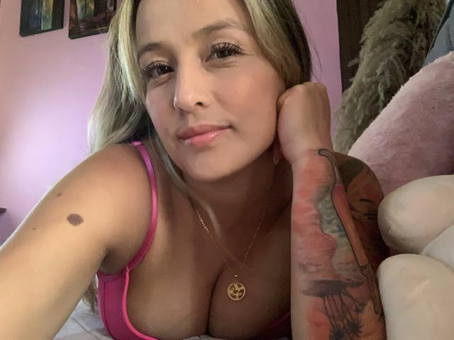 SaritaCaramelo with blonde hair, pink bra, and tattooed arm, smiling at camera, head resting on hand, gold necklace, medium tits, pink room, soft light, relaxed.