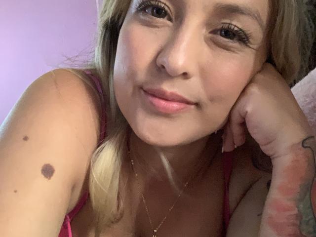 SaritaCaramelo with long blonde hair, light skin, and a small black mole on her right arm, wearing a pink top, smiling at the camera with her hand on her cheek.