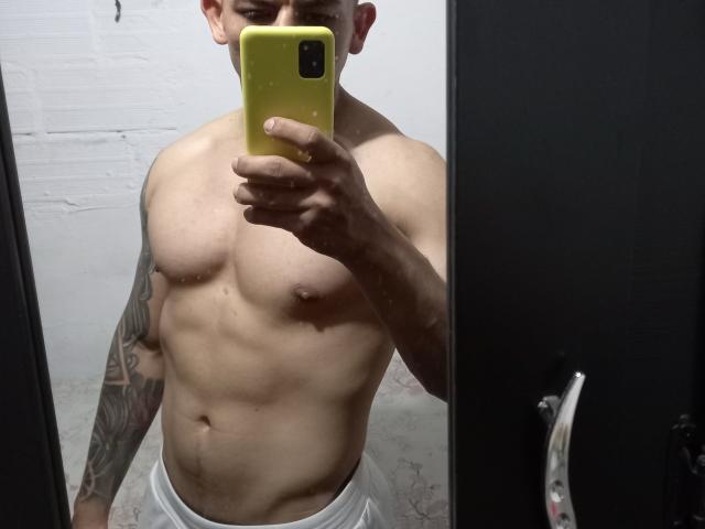 A selfie of DarrinPaul, a muscular, shirtless man with light brown skin and a tattoo on his right arm. He holds a yellow phone in front of his face, covering it. His defined abs and chest are prominent. He wears white pants, and the background is a white brick wall. The image is sharply focused, with a reflective mirror capturing his upper body. The phone's casing is textured. The composition is centered.