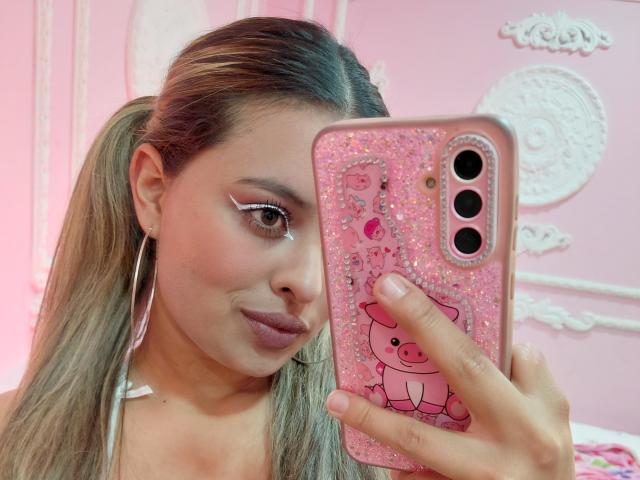 AngellVisen with long blonde hair, wearing pink lipstick, holding a pink glitter phone case with a Hello Kitty sticker, in a pink room. Boobs are not visible.